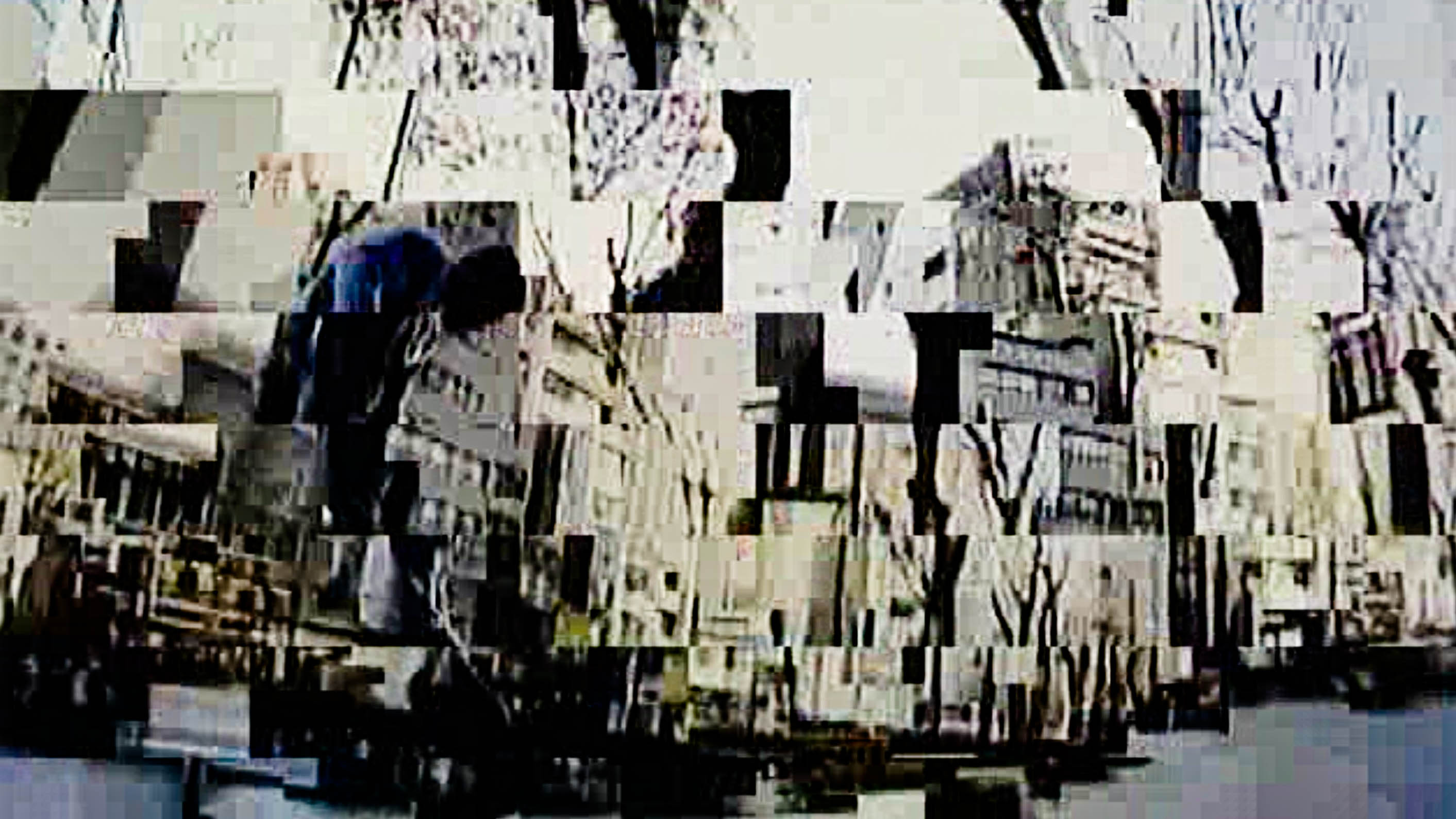 A video still is broken up into a mosaic, with what could be a person hunched over in the left-hand side of the screen. The broken-up background is composed of buildings and trees.
