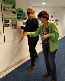 Two people walk the hallway, one person has a cane and sight-restricting glasses.