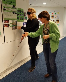 Twee mensen lopen door de gang, één persoon heeft een wandelstok en een zichtbeperkende bril.