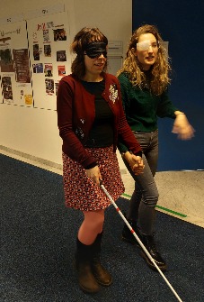 Two people with sight-restricting glasses walk down the hall. The person on the left has a cane for the blind.