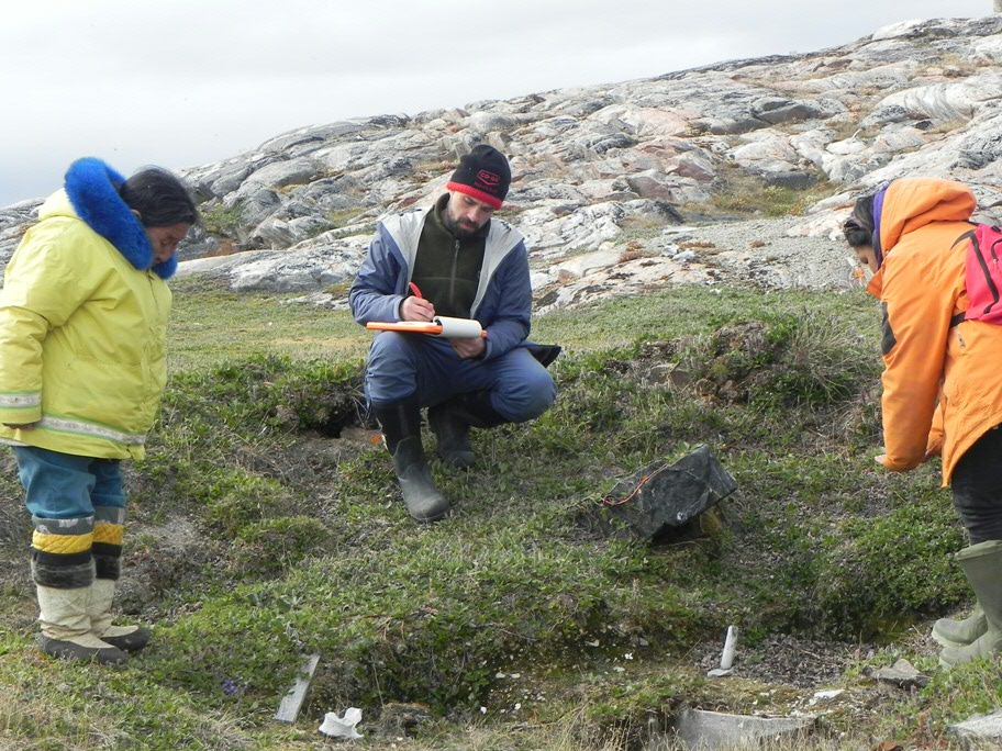 Sean Desjardins (midden) overlegt met Igloolik oudere Madeline Ivalu (links) en Inuktitut vertaler Janet Airut (rechts) bij de overblijkselen van een oud zodenhuis, Avvajja, Nunavut, Canada, 2018. (Photo by Scott Rufolo.)