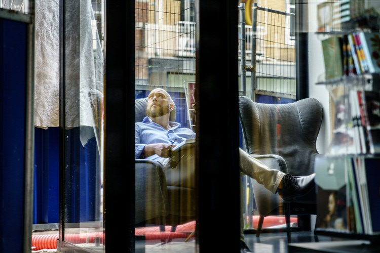 Konstantin Mierau in een oorfauteil achter glas, met een boek en zonnenbril in zijn handen, kijkt achter zich.