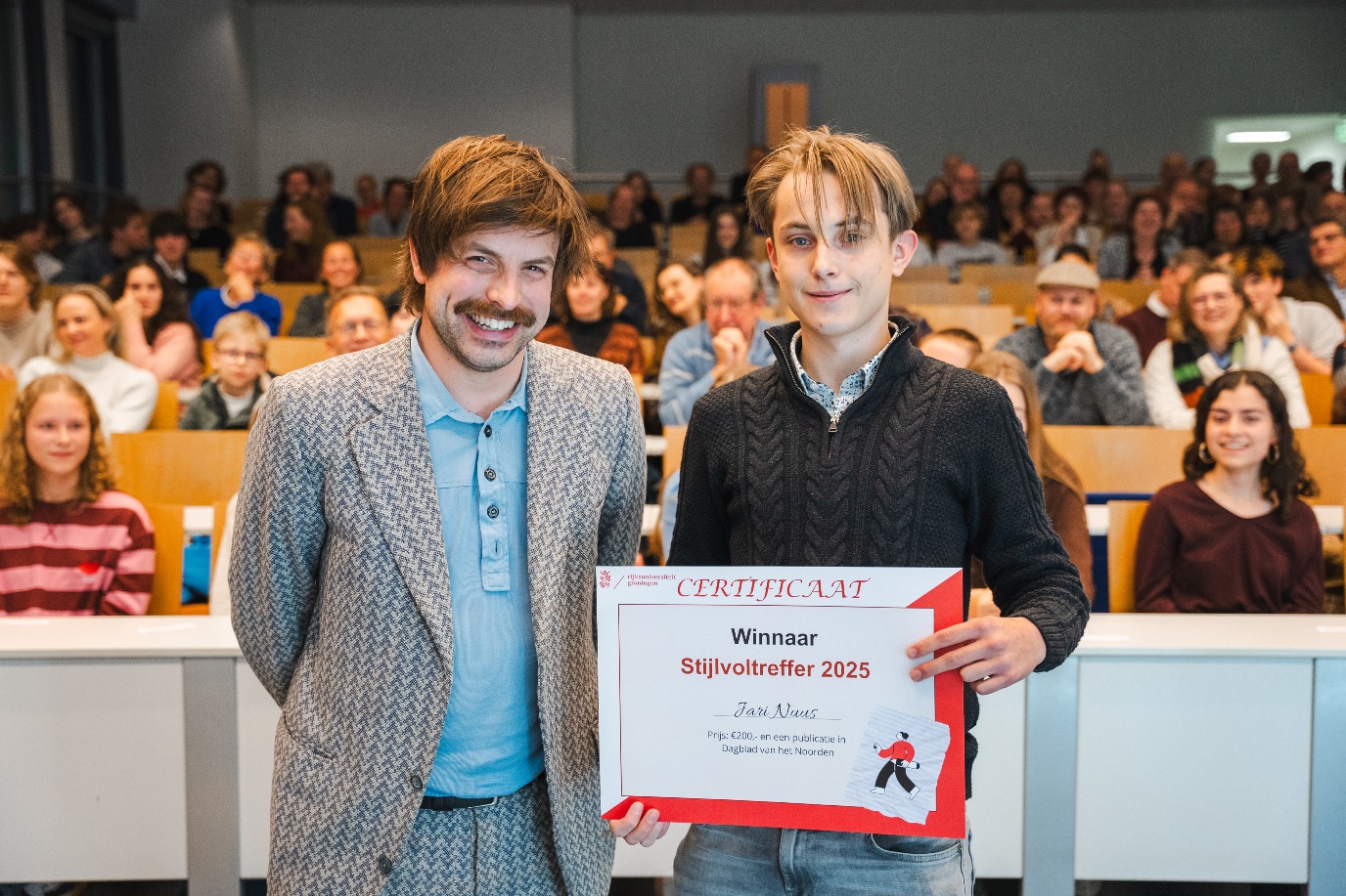 Joost Oomen (links) gastschrijver RUG 2025 - een man met bruin haar en snoor, lachend. Naast hem (rechts) staat een glimlachende jongeman, Jari Nuus, de winnaar van de Stijlvoltreffer 2025. Hij houdt een prijzengeldcheque vast. Ze staan voor een zaal vol studenten en andere gasten.