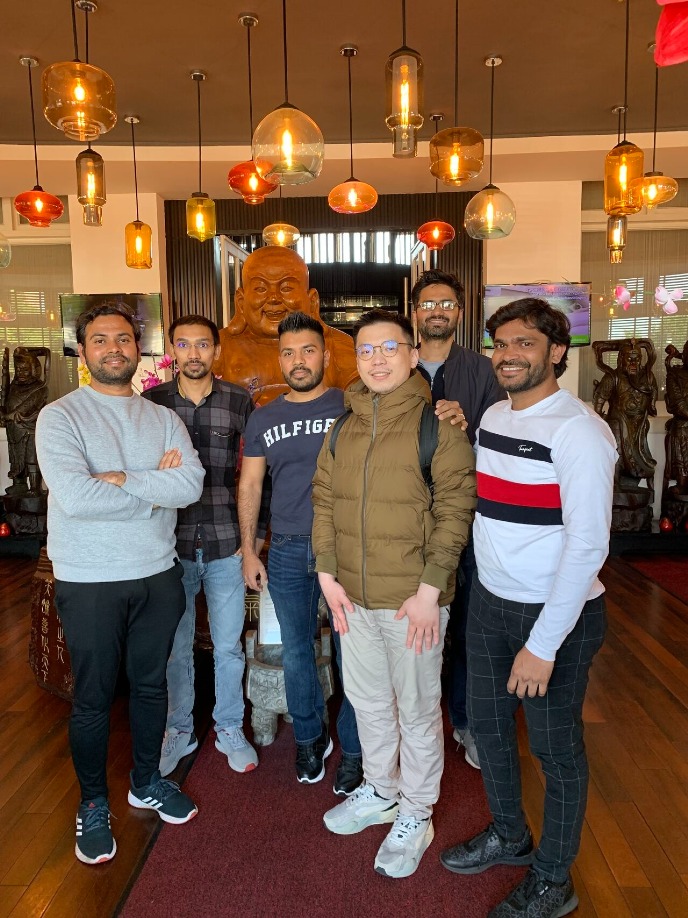 The group members and the chairman are in a hall of a restaurant under a bunch of colourful lights hanging from the ceiling