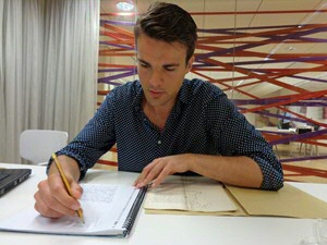 Bram van Leuveren takes notes in a spiral-bound notebook. To its left lies a handwritten archival document on its typical tan folder. In the background a clear glass wall is criss-crossed by multicoloured lines.