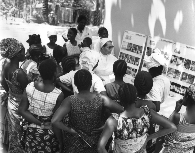 A white woman in a white habit and veil stands in front of three large posters with images, closely surrounded by a group of young black women, on of which carries a baby on her hip.