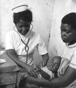 A young black woman holds a baby on her lap as a black nurse puts a stethoscope to the baby's chest.