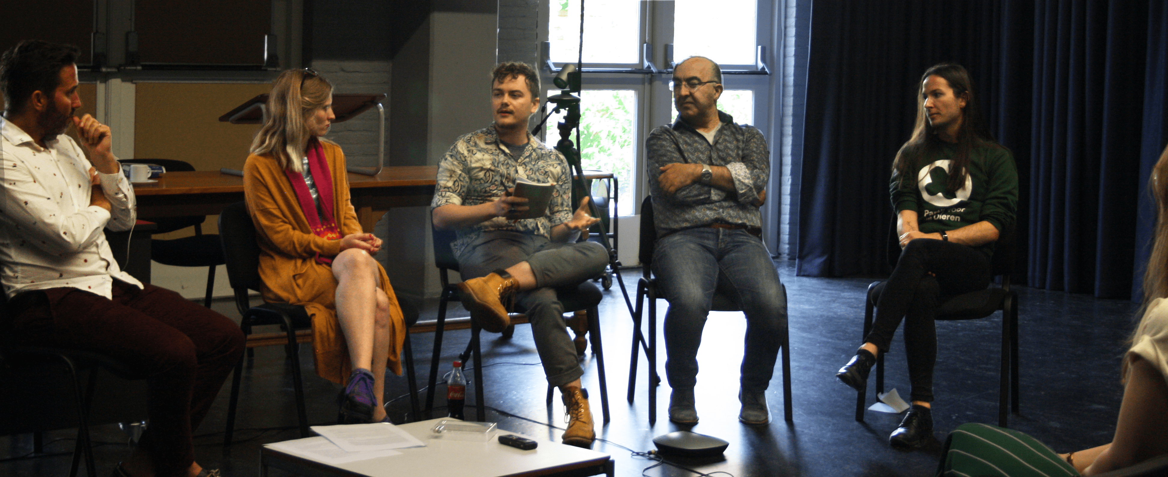 A photo of the roundtable discussion: six people are sitting on chairs, five are facing the viewer, one is with their back to the camera. One is speaking while holding a notebook, the others are listening attentively.