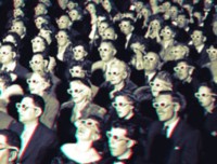 A black and white photograph of a theatre filled with neatly-dressed white people looking at something (presumably a movie screen) while wearing bicolored 3D-glasses.