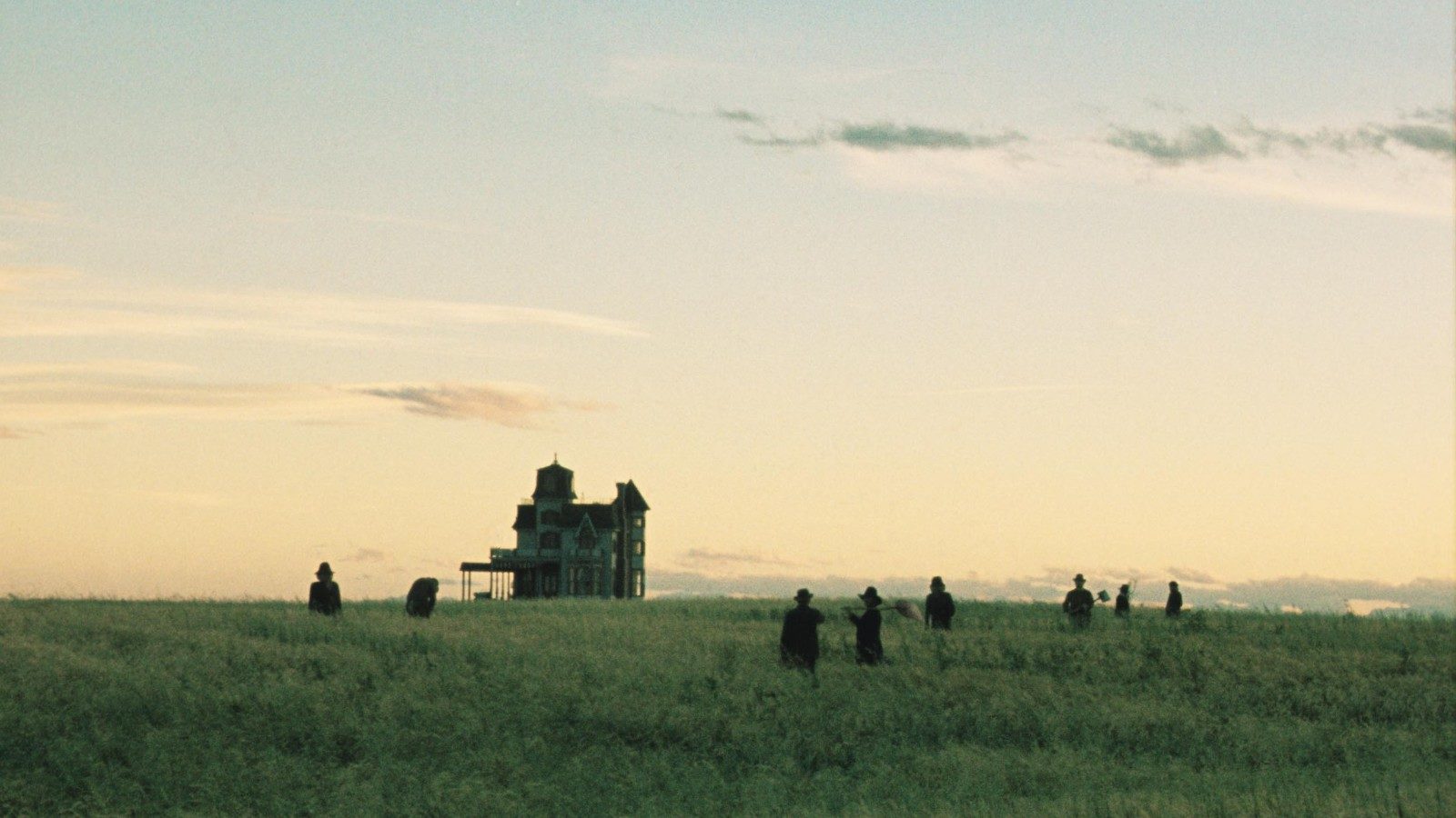 Still from the movie Days of Heaven (1978). The color image shows the silhouets of 8 people spread out in a field, and a tall house in the distance, under en open sky.