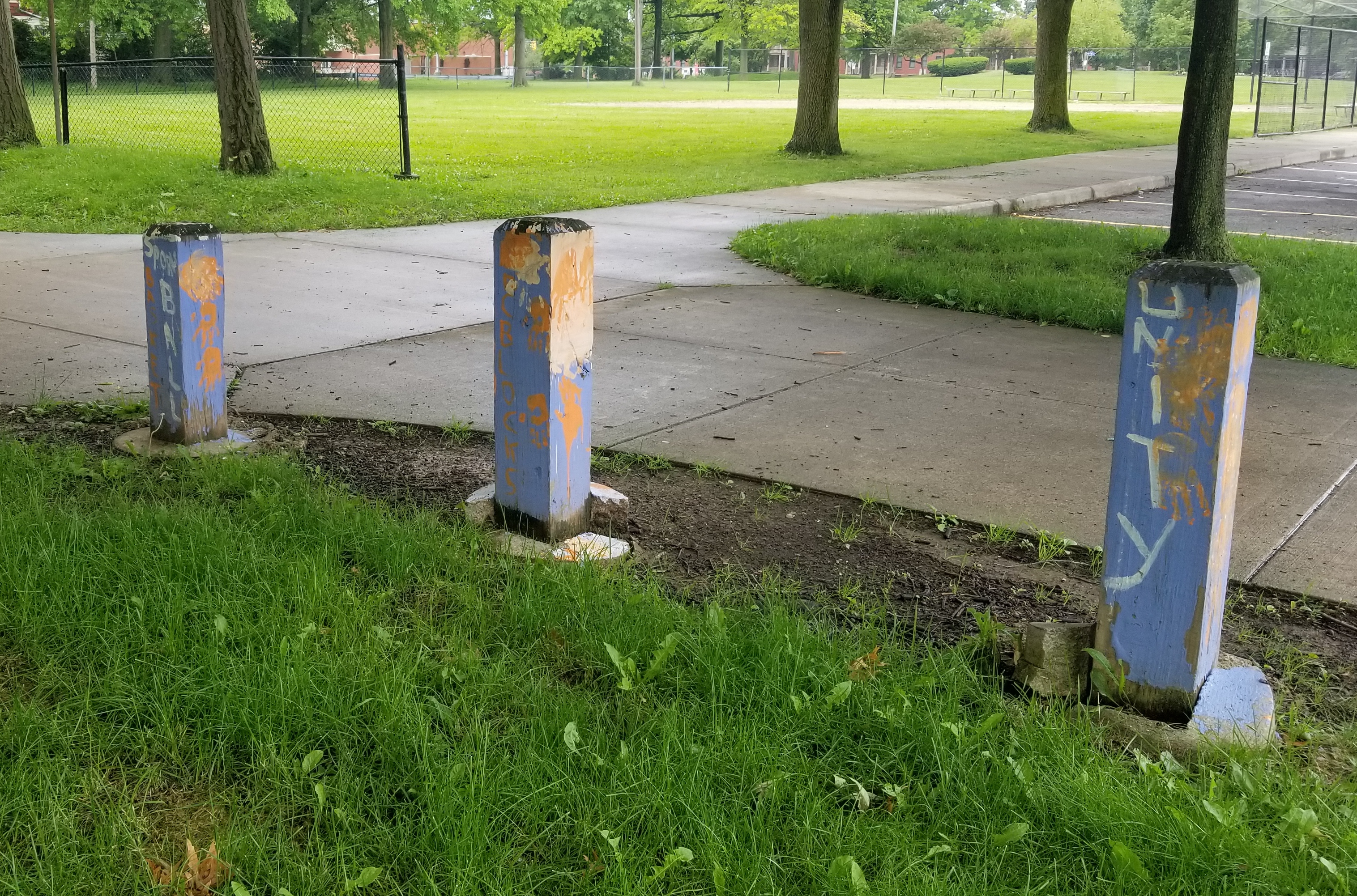Three short square concrete poles along a path in a park. They are pained light blue with orange hand prints and white text spelling, among other things, "unity".