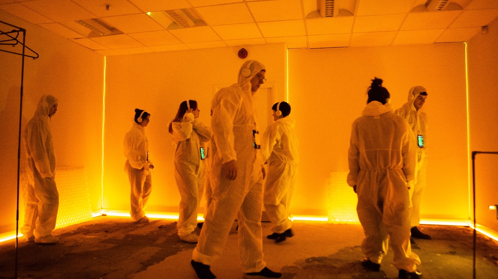 A color photograph of seven persons walking around in white disposable overalls and headphones in an orange-lit windowless room.