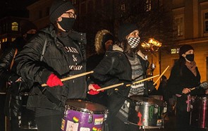 photo showing three protesters from the group Rhythms of Resistance Wrocław drumming in the street. The protesters are masked and dressed in black; the drums have stickers on them, which include rainbow flags.
