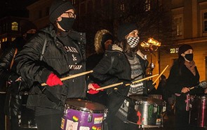 Zdjęcie przedstawiające trzech protestujących z grupy Rytmy Oporu Wrocław bębniących na ulicy. Protestujący są zamaskowani i ubrani na czarno; na bębnach mają naklejki, wśród których widnieją tęczowe flagi.